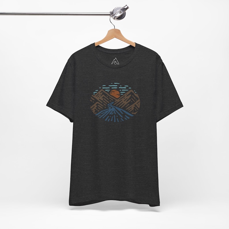 May include: A dark heather grey t-shirt with a graphic of a mountain range with a sun setting behind it. The graphic is in shades of brown, blue, and orange.