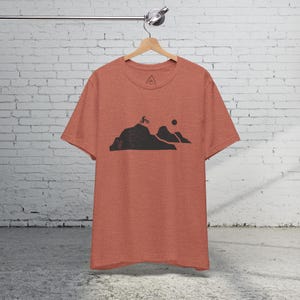 K&ouml;nnte beinhalten: Ein rotes Melange-T-Shirt mit einer schwarzen Grafik eines Mountainbikers, der &uuml;ber einen H&uuml;gel f&auml;hrt, mit einer Sonne im Hintergrund.