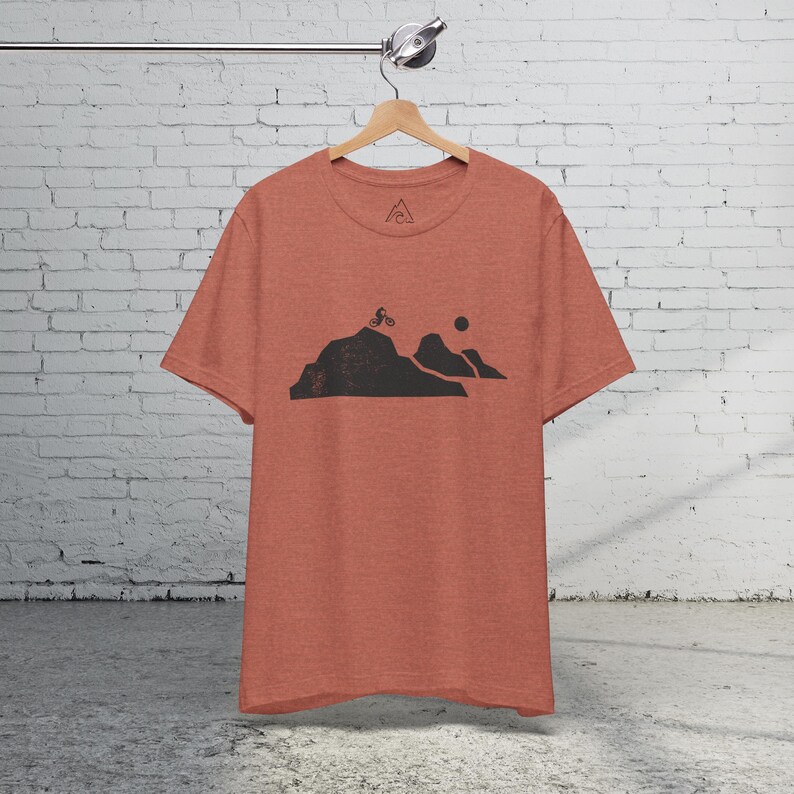 K&ouml;nnte beinhalten: Ein rotes Melange-T-Shirt mit einer schwarzen Grafik eines Mountainbikers, der &uuml;ber einen H&uuml;gel f&auml;hrt, mit einer Sonne im Hintergrund.