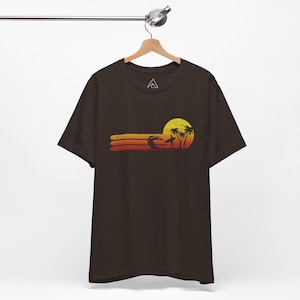 May include: A dark brown t-shirt with a retro sunset graphic featuring a surfer riding a wave, palm trees, and a yellow sun. The graphic is in shades of orange, yellow, and red.