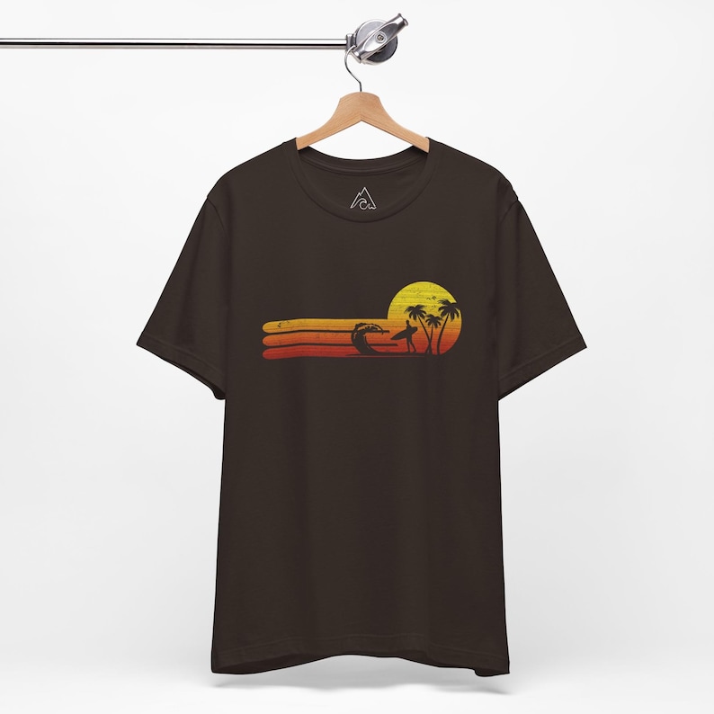 May include: A dark brown t-shirt with a retro sunset graphic featuring a surfer riding a wave, palm trees, and a yellow sun. The graphic is in shades of orange, yellow, and red.