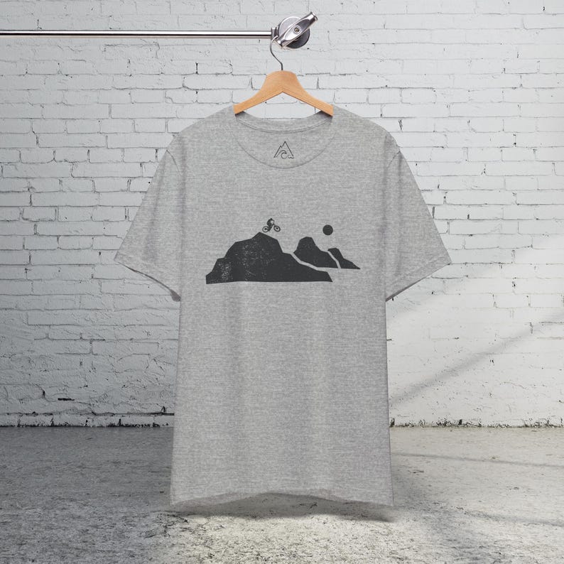 K&ouml;nnte beinhalten: Ein hellgraues T-Shirt mit einem Aufdruck eines Mountainbikers, der einen Berg hinauf f&auml;hrt, mit einer Sonne im Hintergrund.