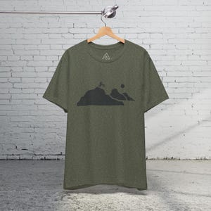K&ouml;nnte beinhalten: Ein gr&uuml;nes T-Shirt mit einer schwarzen Silhouette einer Bergkette und einer Person, die mit dem Fahrrad einen H&uuml;gel hinauf f&auml;hrt. Das T-Shirt hat ein kleines Logo auf der Vorderseite.
