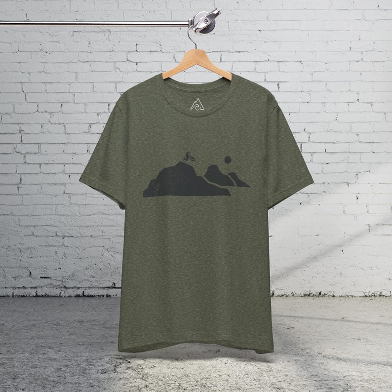 K&ouml;nnte beinhalten: Ein gr&uuml;nes T-Shirt mit einer schwarzen Silhouette einer Bergkette und einer Person, die mit dem Fahrrad einen H&uuml;gel hinauf f&auml;hrt. Das T-Shirt hat ein kleines Logo auf der Vorderseite.