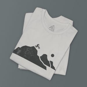 K&ouml;nnte beinhalten: Wei&szlig;es T-Shirt mit einer schwarzen Grafik eines Mountainbikers, der auf einem Bergpfad f&auml;hrt. Die Grafik ist im Vintage-Stil mit einem Used-Look. Das T-Shirt hat ein kleines schwarzes Logo auf der Vorderseite, das "AEROSOL" sagt.