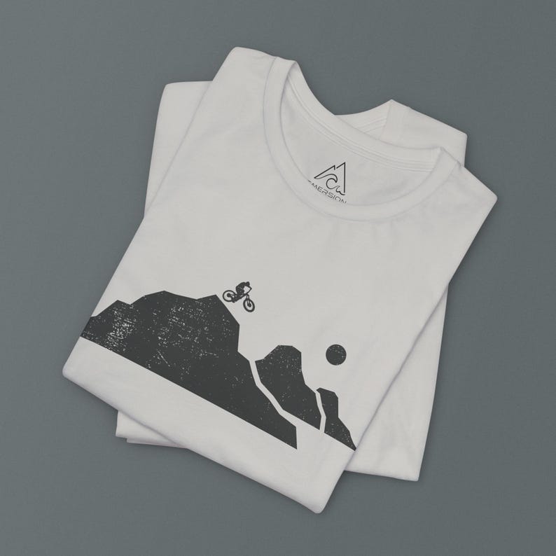 K&ouml;nnte beinhalten: Wei&szlig;es T-Shirt mit einer schwarzen Grafik eines Mountainbikers, der auf einem Bergpfad f&auml;hrt. Die Grafik ist im Vintage-Stil mit einem Used-Look. Das T-Shirt hat ein kleines schwarzes Logo auf der Vorderseite, das "AEROSOL" sagt.