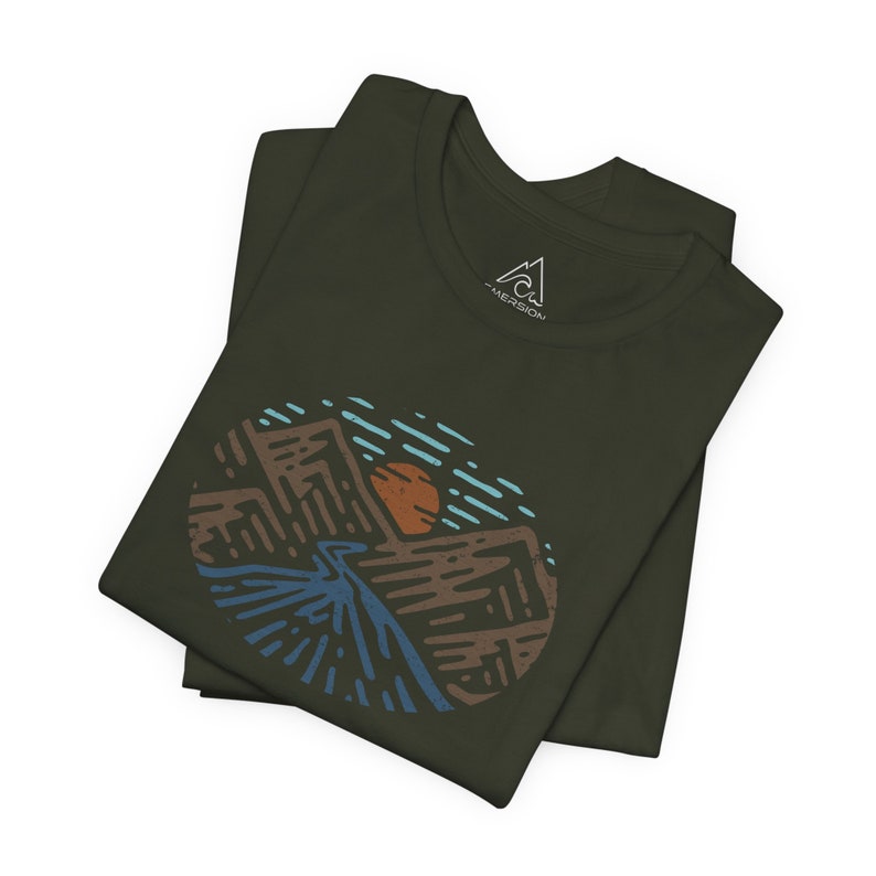 May include: A folded green t-shirt with a circular graphic of a mountain range, a river, and a sun. The graphic is in shades of brown, blue, and orange. The t-shirt has the word "MERISON" printed on the collar.