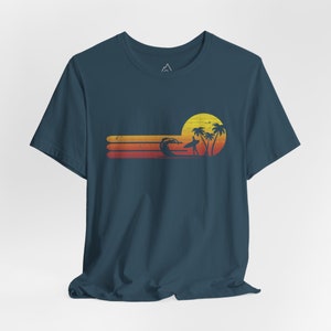 May include: A dark blue t-shirt with a retro sunset graphic featuring a surfer, palm trees, and a yellow sun. The graphic is in shades of orange, yellow, and red.