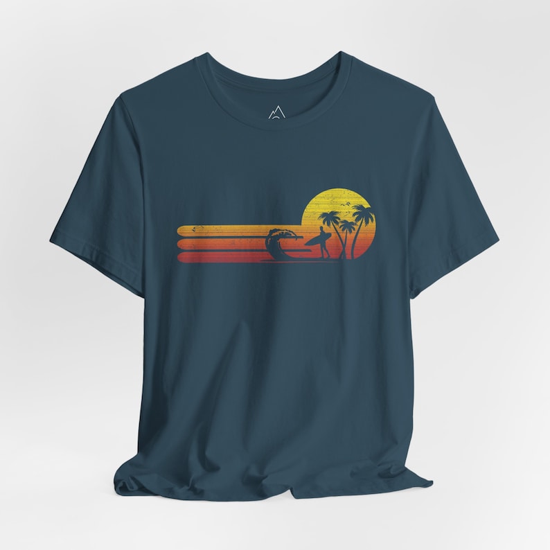 May include: A dark blue t-shirt with a retro sunset graphic featuring a surfer, palm trees, and a yellow sun. The graphic is in shades of orange, yellow, and red.