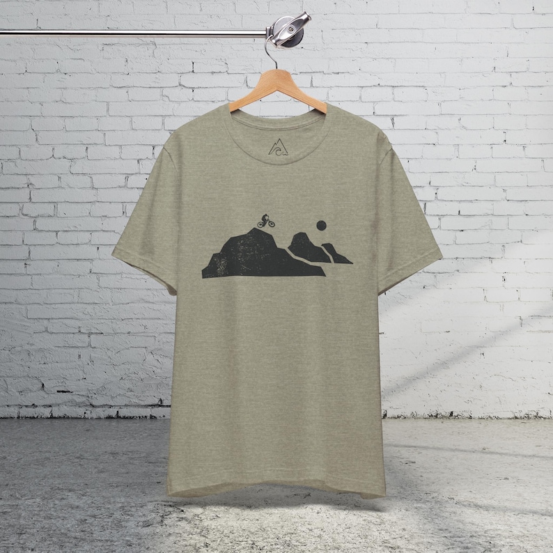 K&ouml;nnte beinhalten: Ein hellgraues T-Shirt mit einer schwarzen Grafik eines Mountainbikers, der einen H&uuml;gel mit einer Sonne im Hintergrund hinauf f&auml;hrt.
