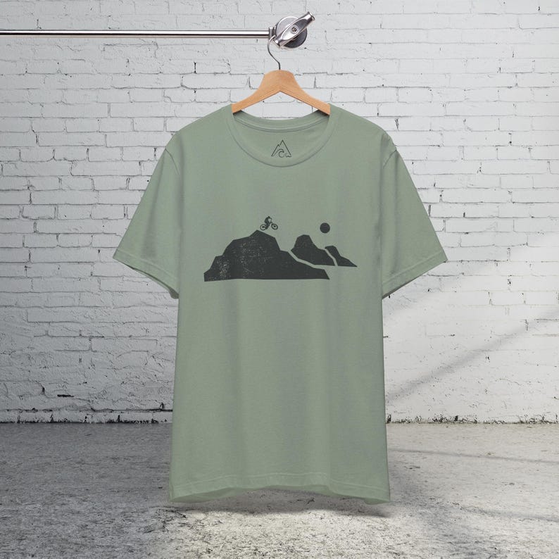 K&ouml;nnte beinhalten: Ein salbeigr&uuml;nes T-Shirt mit einer schwarzen Grafik eines Mountainbikers, der einen H&uuml;gel mit einer Sonne im Hintergrund hinauf f&auml;hrt.