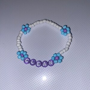 May include: A beaded bracelet with white, light blue, and purple beads. The bracelet features light blue flower accents and the word "CLEAN" spelled out in purple letter beads.