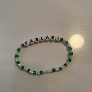 May include: A beaded bracelet spelling "PABLASI" with white letter beads. The bracelet has alternating green and silver beads. The design is simple and elegant. The bracelet is likely handmade.