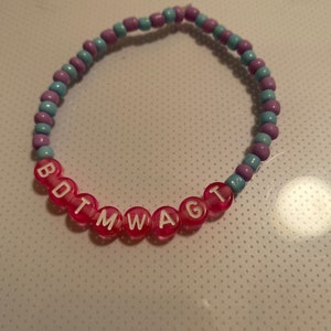 Può includere: Un braccialetto di perline con perline rosa, azzurre e viola. Il braccialetto ha perline con lettere rosa trasparenti che compongono "BOTMWAGT". Il braccialetto è su una superficie bianca.