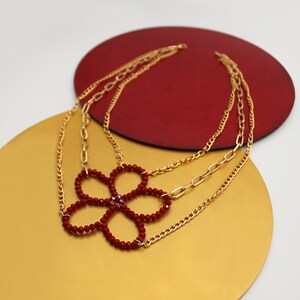 Peut inclure: Un collier en chaîne dorée avec un pendentif en forme de fleur rouge fait de petites perles. La fleur a un petit strass transparent au centre.