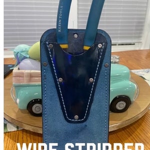 May include: A blue leather wire stripper pocket with a blue and white stitched pocket and two blue wire strippers with yellow handles. The strippers have the text "KLEIN TOOLS" printed on them. The pocket is labeled "WIRE STRIPPER POCKET".