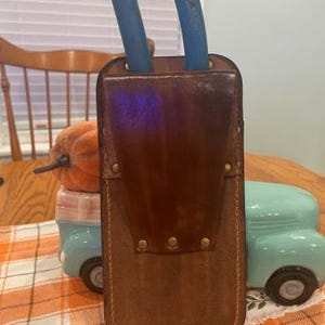May include: A brown leather tool holder with two blue pruning shears inserted. The holder has a belt loop and brass rivets.