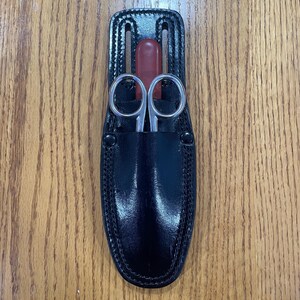 May include: A black leather sheath with a stitched edge holds a pair of silver scissors and a red tool.