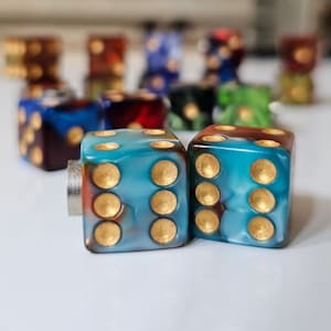 May include: Two blue dice with gold dots, one with a brown swirl and the other with a red swirl. The dice are on a white surface.