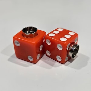 May include: Two red dice with white dots and metal rings. The dice are cube-shaped and have the traditional dot patterns. The metal rings are on top and on the side of the dice.
