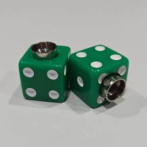 May include: Two green dice with white dots and metal fittings. The dice are cube-shaped and have a shiny, reflective surface. The white dots indicate the numbers on each die. The metal fittings are cylindrical and are set into the top of each die.