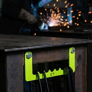 May include: A neon green clamp holder attached to a metal table with bolts. The holder has a series of slots to hold clamps. The text "GY" is visible on the holder. The image also includes the text "A home for your clamps".