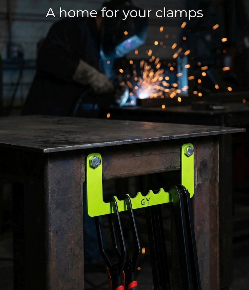 May include: A neon green clamp holder attached to a metal table with bolts. The holder has a series of slots to hold clamps. The text "GY" is visible on the holder. The image also includes the text "A home for your clamps".