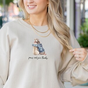 May include: A beige sweatshirt featuring a graphic of a dog and a person embracing. The text "paws and give thanks" is printed beneath the graphic.