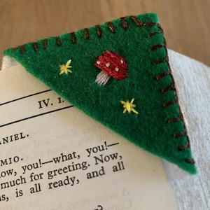 Può includere: Un segnalibro in feltro verde con un bordo cucito marrone. Il segnalibro ha la forma di un triangolo e presenta un fungo rosso e bianco ricamato con stelle gialle.