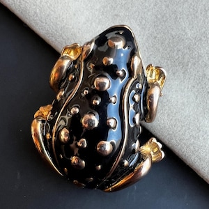 Peut inclure: Une broche en forme de grenouille noire et dorée avec une surface texturée. La grenouille est posée sur une surface gris clair.