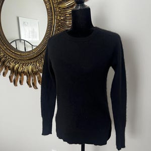 May include: A black crew neck sweater displayed on a black mannequin. The sweater has long sleeves and a slightly rounded hem. The background includes a gold sunburst mirror and a white wall.