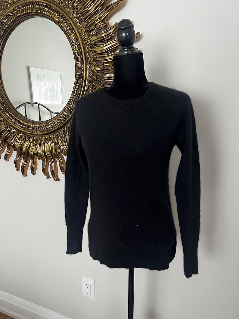 May include: A black crew neck sweater displayed on a black mannequin. The sweater has long sleeves and a slightly rounded hem. The background includes a gold sunburst mirror and a white wall.