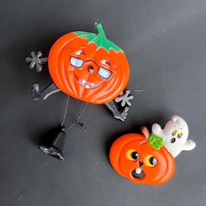 May include: Two Halloween-themed pumpkin pins. One is orange with a smiling face, wearing glasses and has black arms and legs. The other is orange with a ghost on top and has a black mouth and eyes.