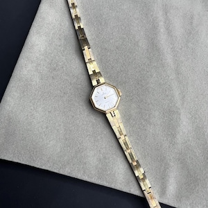 May include: A gold-coloured wristwatch with a white face and an octagonal bezel. The watch has a delicate, linked strap. The watch is displayed on a grey surface.