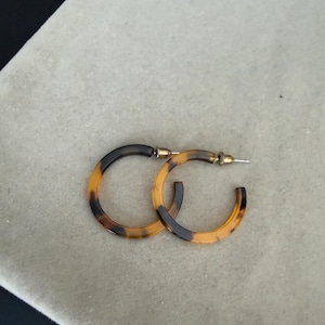 Op de afbeelding: Een paar oorringen met een bruin en zwart schildpadpatroon. De oorbellen zijn cirkelvormig en hebben een metalen stift-sluiting. Het ontwerp heeft een gladde, gepolijste afwerking over de gehele oppervlakte van de oorringen.