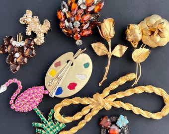 May include: A collection of vintage brooches featuring a variety of designs, including a rose, a paint palette, a flamingo, a bear, and a leaf. The brooches are made of gold metal and rhinestones.