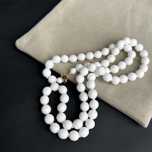 May include: A white beaded necklace with a gold clasp, resting on a beige fabric. The necklace is made of many round, white beads. The background is a dark surface.