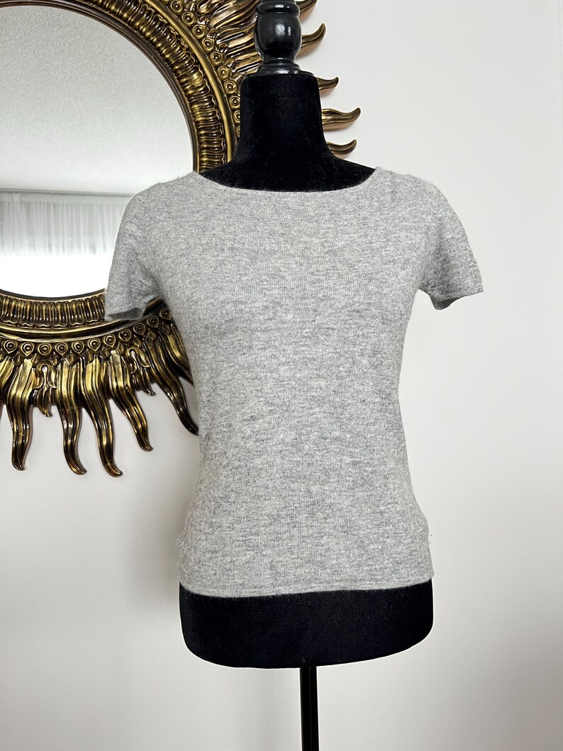 K&ouml;nnte beinhalten: Ein meliertes, graues Kurzarmshirt mit Bootsausschnitt. Das Shirt wird auf einer schwarzen Schaufensterpuppe pr&auml;sentiert. Im Hintergrund ein goldgerahmter Spiegel.
