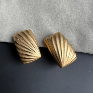 May include: A pair of gold-toned metal earrings with a textured, fan-like shell pattern. The earrings have a rectangular, slightly curved shape with a matte finish and radiating grooves that extend from one corner across the surface.