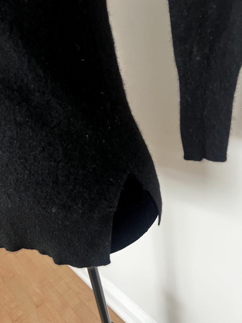May include: A black sweater with a split hem and long sleeves. The sweater is made of a soft, textured material. The hemline is slightly curved, and the sleeves are long and fitted.