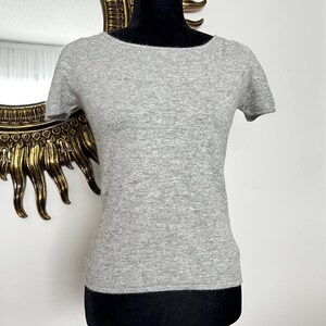 K&ouml;nnte beinhalten: Ein hellgraues Kurzarm-Oberteil mit rundem Ausschnitt. Das Shirt besteht aus einem weichen, strukturierten Material und wird auf einer schwarzen Schaufensterpuppe pr&auml;sentiert. Der Hintergrund umfasst einen goldgerahmten Spiegel.