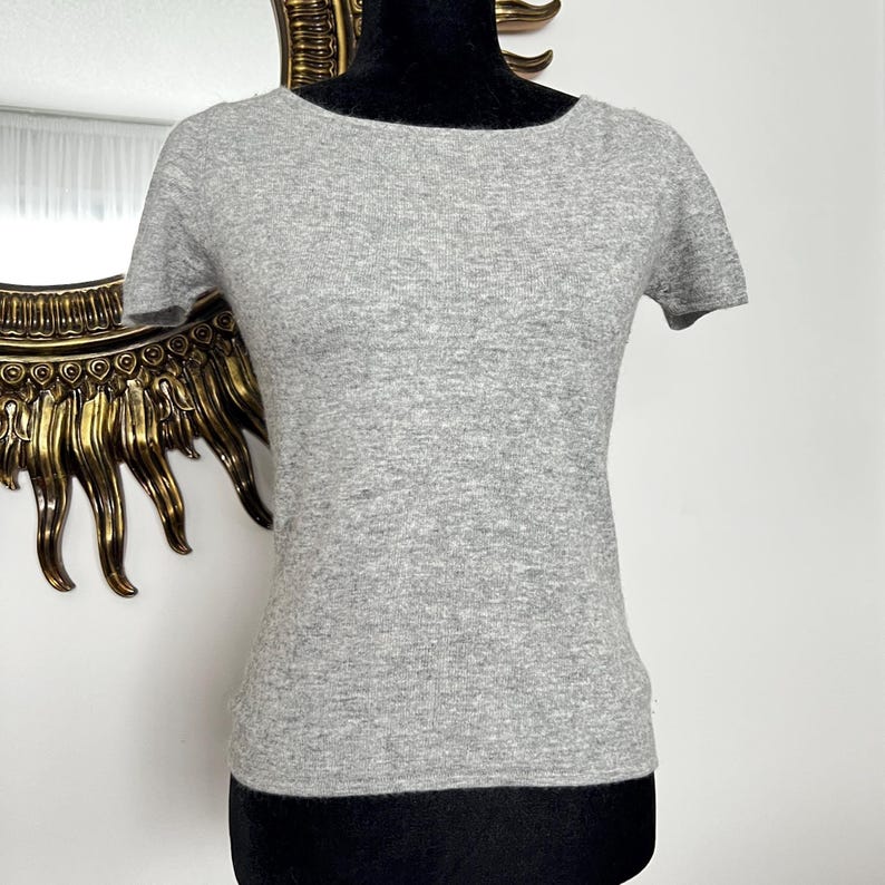 K&ouml;nnte beinhalten: Ein hellgraues Kurzarm-Oberteil mit rundem Ausschnitt. Das Shirt besteht aus einem weichen, strukturierten Material und wird auf einer schwarzen Schaufensterpuppe pr&auml;sentiert. Der Hintergrund umfasst einen goldgerahmten Spiegel.