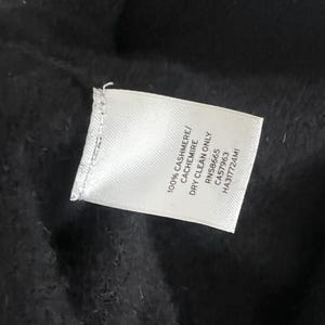 May include: A close-up of a white clothing tag with black text, lying on a black fabric. The tag reads "100% CASHMERE/ CACHEMIRE DRY CLEAN ONLY RN58665 CA57963 HA317724MI".