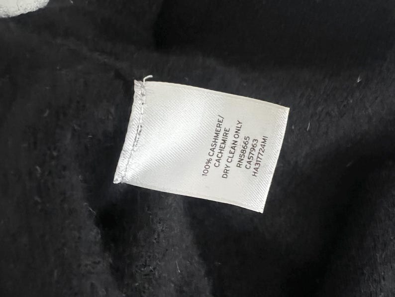 May include: A close-up of a white clothing tag with black text, lying on a black fabric. The tag reads "100% CASHMERE/ CACHEMIRE DRY CLEAN ONLY RN58665 CA57963 HA317724MI".