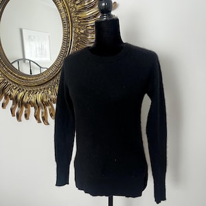 May include: A black crew neck sweater displayed on a mannequin. The sweater has long sleeves and a slightly rounded hemline. The background includes a gold-framed mirror and a white wall.