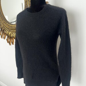May include: A black crew neck sweater with long sleeves. The sweater is displayed on a black mannequin. The sweater has a soft, textured appearance and a classic, versatile design. The background includes a gold-framed mirror.