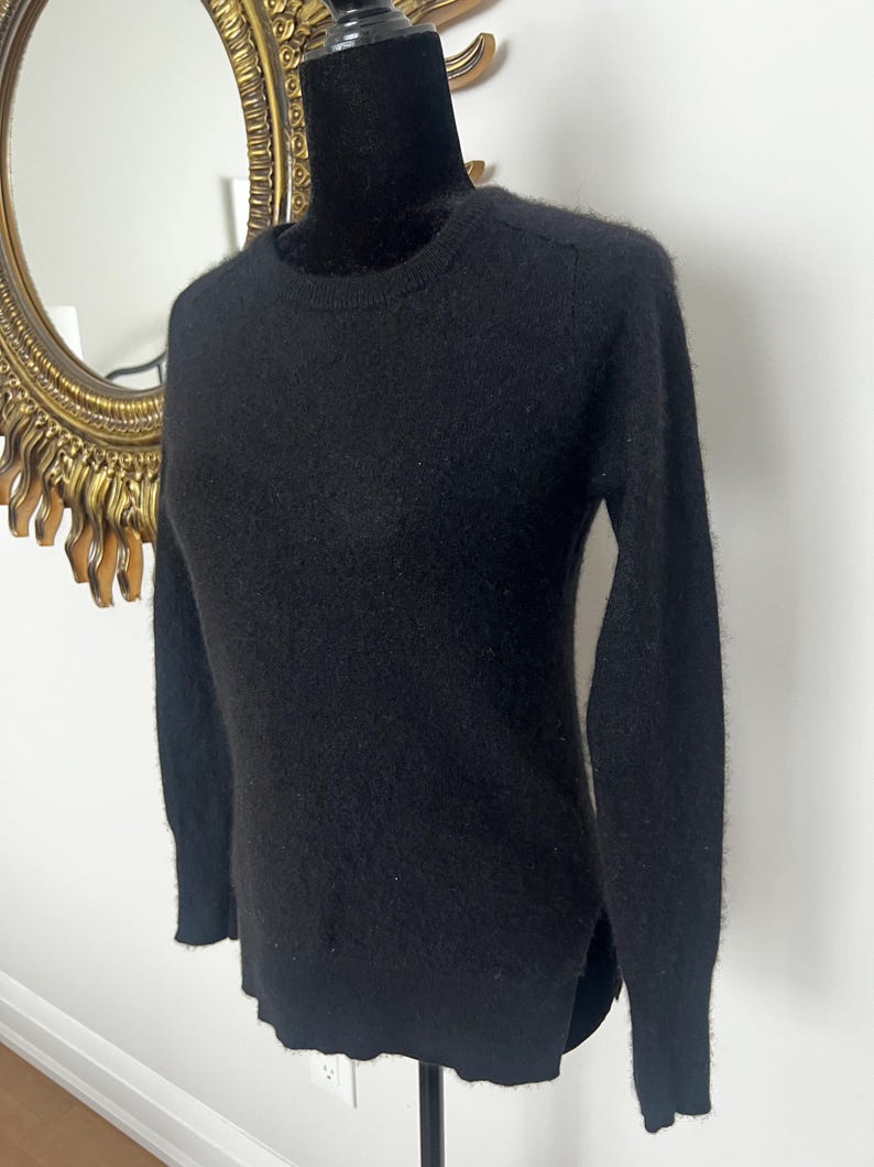 May include: A black crew neck sweater with long sleeves. The sweater is displayed on a black mannequin. The sweater has a soft, textured appearance and a classic, versatile design. The background includes a gold-framed mirror.