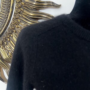 May include: Close-up of a black sweater with a high neckline. The sweater is made of a soft, possibly knitted material. The image is well-lit, highlighting the texture and details of the garment. The background includes a decorative gold-colored mirror.