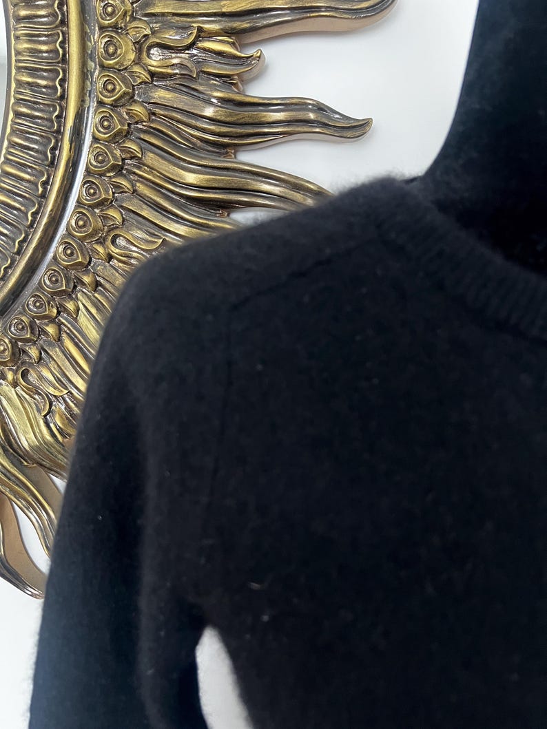 May include: Close-up of a black sweater with a high neckline. The sweater is made of a soft, possibly knitted material. The image is well-lit, highlighting the texture and details of the garment. The background includes a decorative gold-colored mirror.