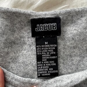 K&ouml;nnte beinhalten: Nahaufnahme eines grauen Pullovers mit einem schwarzen Etikett. Das Etikett zeigt "JACOB" in wei&szlig;en Blockbuchstaben. Darunter die Gr&ouml;&szlig;e "M" und eine Liste der Stoffe, darunter Viskose, Nylon, Wolle und Angora-Kaninchenhaar. Hergestellt in Hongkong.
